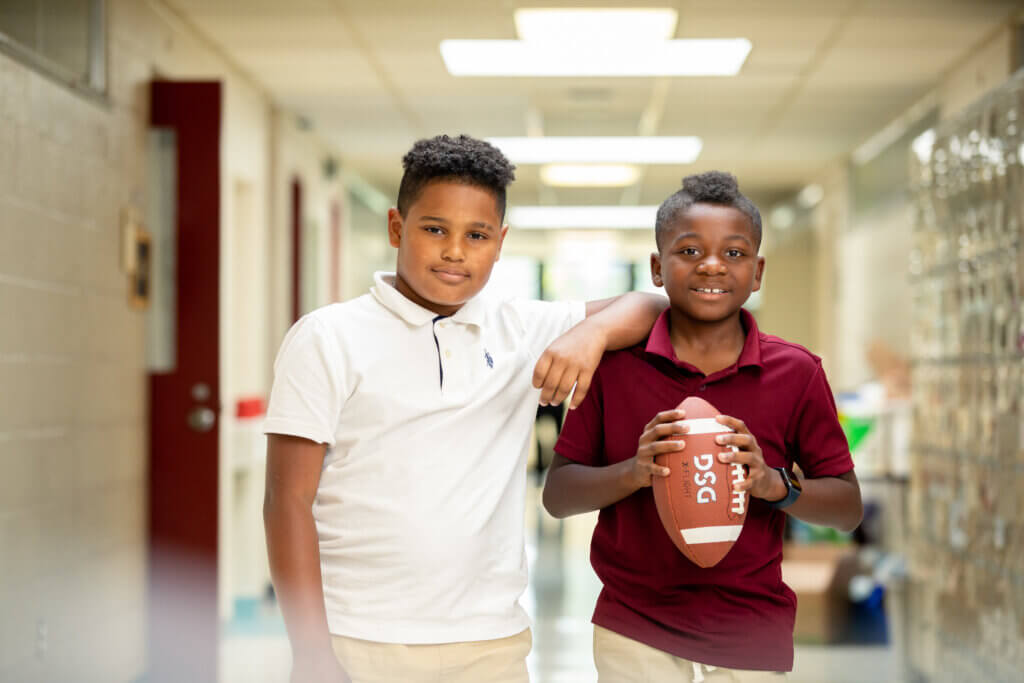 Photo of 2 Four Corners Montessori Academy students posing for the camera in the school hallway, one holding a football and both smiling.